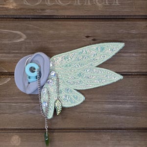 May include: A light blue earplug with a silver chain attachment featuring a green leaf charm and clear rhinestones. Attached to the chain is a large, iridescent, light green fabric wing ornament with a detailed, textured, vein-like pattern.