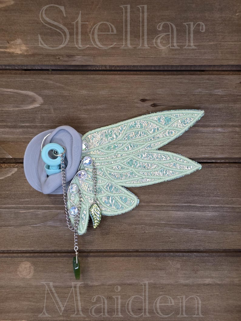 May include: A light blue earplug with a silver chain attachment featuring a green leaf charm and clear rhinestones. Attached to the chain is a large, iridescent, light green fabric wing ornament with a detailed, textured, vein-like pattern.