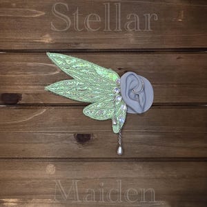 May include: An ear cuff featuring iridescent light green fairy wings with intricate silver vein detailing. The piece includes a silver chain, small round iridescent beads, and a teardrop-shaped charm. The word Stellar is above and Maiden is below.