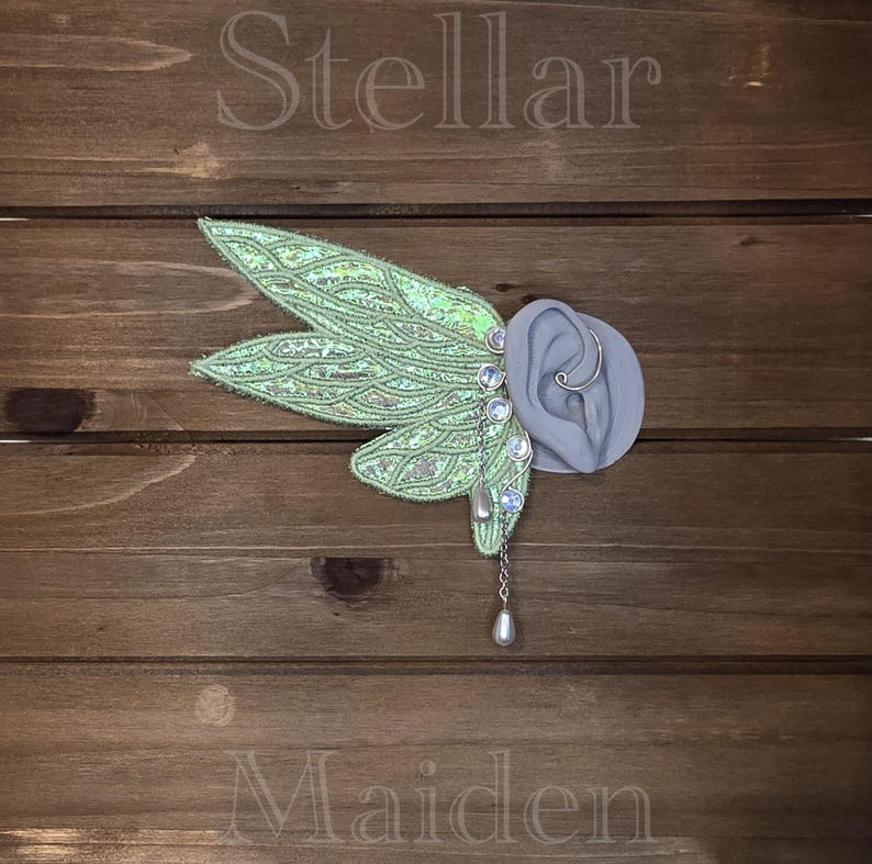 May include: An ear cuff featuring iridescent light green fairy wings with intricate silver vein detailing. The piece includes a silver chain, small round iridescent beads, and a teardrop-shaped charm. The word Stellar is above and Maiden is below.