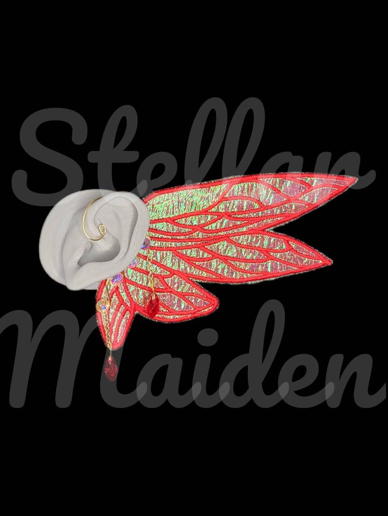 May include: An ear cuff featuring iridescent red wings with gold-toned veins and dangling red crystal accents. The accessory is displayed on a neutral-toned ear model. The text "Stellar Maiden" is overlaid in a dark, semi-transparent script font.