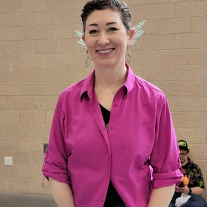 May include: A bright magenta button-down shirt with a collar and rolled-up sleeves, worn with silver-toned dangling earrings featuring circular wire frames and translucent, pale green wing-shaped ear cuffs attached to the upper ear cartilage.
