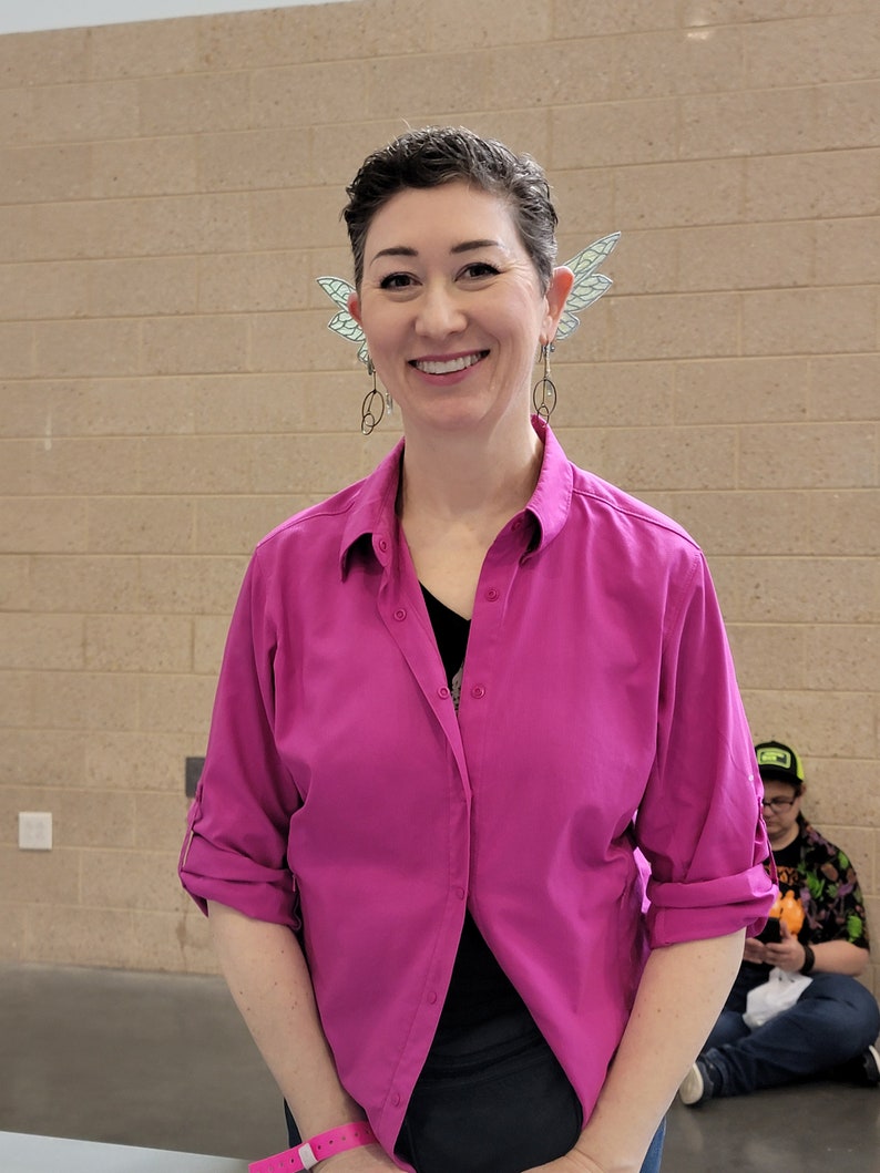 May include: A bright magenta button-down shirt with a collar and rolled-up sleeves, worn with silver-toned dangling earrings featuring circular wire frames and translucent, pale green wing-shaped ear cuffs attached to the upper ear cartilage.