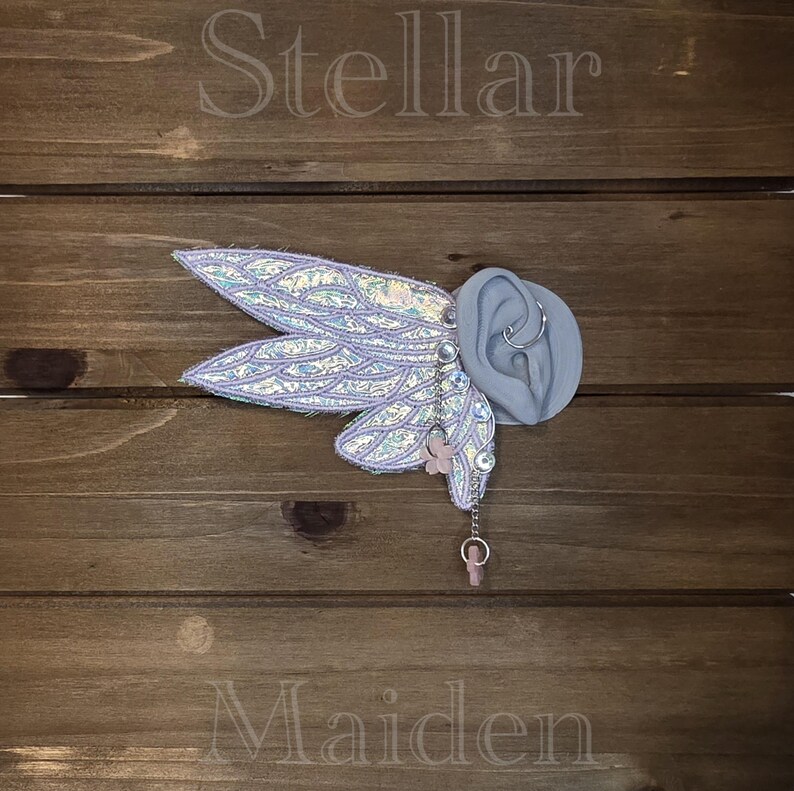 May include: An ear cuff featuring iridescent, light purple, wing-shaped fabric attachments with a scale-like pattern. The piece includes a silver-toned chain, clear rhinestones, and a small pink flower charm, all displayed on a gray ear model.