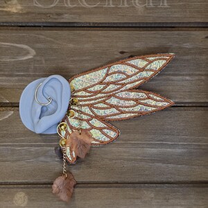 May include: An ear cuff featuring iridescent, wing-shaped panels with copper-colored stitching and veins. The piece includes gold-toned circular accents, a delicate chain, and two translucent, light brown leaf-shaped charms attached to the structure.