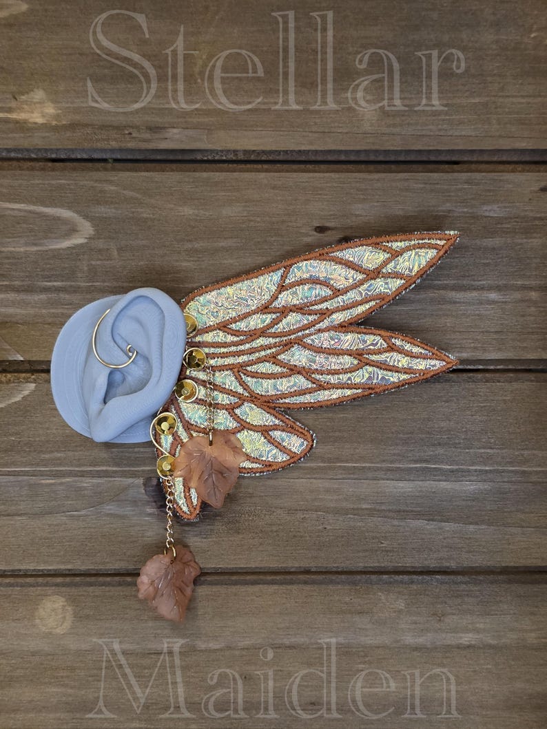 May include: An ear cuff featuring iridescent, wing-shaped panels with copper-colored stitching and veins. The piece includes gold-toned circular accents, a delicate chain, and two translucent, light brown leaf-shaped charms attached to the structure.