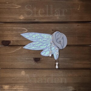 May include: An ear cuff featuring iridescent, light blue, wing-shaped fabric attachments with silver-toned wire detailing. The piece includes small white pearl beads and dangling teardrop-shaped charms. The word Stellar is above and Maiden is below.