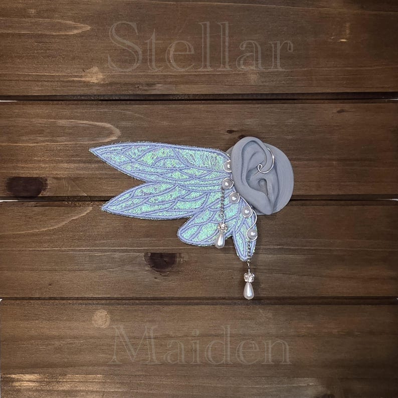 May include: An ear cuff featuring iridescent, light blue, wing-shaped fabric attachments with silver-toned wire detailing. The piece includes small white pearl beads and dangling teardrop-shaped charms. The word Stellar is above and Maiden is below.
