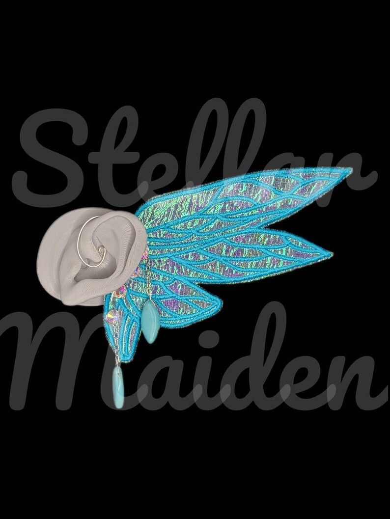 May include: A light gray ear-shaped display form featuring a silver hoop earring and a large, iridescent blue and purple wing-shaped attachment. Two dangling charms, one clear faceted bead and one smooth turquoise teardrop, hang from the wing.