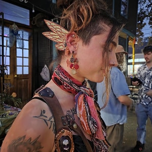May include: A gold-toned ear cuff featuring two layered, glittery wing-shaped attachments. The accessory is worn on the upper ear cartilage, complemented by a dangling earring with red, orange, and gold beads and a circular charm hanging below the lobe.