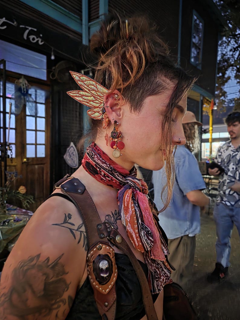 May include: A gold-toned ear cuff featuring two layered, glittery wing-shaped attachments. The accessory is worn on the upper ear cartilage, complemented by a dangling earring with red, orange, and gold beads and a circular charm hanging below the lobe.