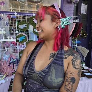 May include: A pair of iridescent teal and black butterfly wing ear cuffs featuring intricate vein patterns and small dangling silver chains, worn on the side of the ear, showcasing a shimmering finish that reflects light with a metallic sheen.
