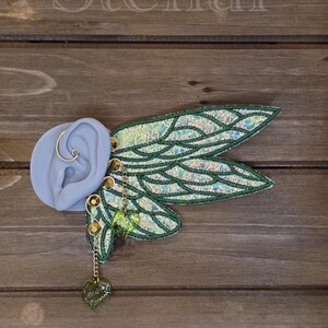 May include: An ear cuff featuring iridescent, wing-shaped fabric attachments with dark green embroidered veins. The piece includes gold-toned metal accents, a thin wire loop, and two dangling, translucent green leaf-shaped charms. The text Stellar Maiden is visible.