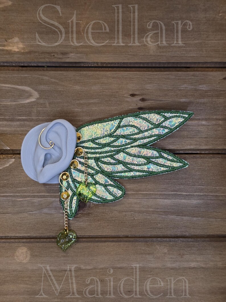 May include: An ear cuff featuring iridescent, wing-shaped fabric attachments with dark green embroidered veins. The piece includes gold-toned metal accents, a thin wire loop, and two dangling, translucent green leaf-shaped charms. The text Stellar Maiden is visible.