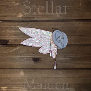 May include: An ear cuff featuring iridescent, wing-shaped fabric attachments with pink trim. The design includes dangling gold-tone chains adorned with small white pearls and purple beads. The word Stellar is printed above and Maiden is printed below.