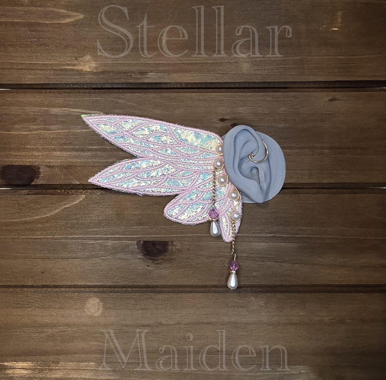 May include: An ear cuff featuring iridescent, wing-shaped fabric attachments with pink trim. The design includes dangling gold-tone chains adorned with small white pearls and purple beads. The word Stellar is printed above and Maiden is printed below.