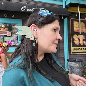 May include: A black headband featuring a layered blue flower ornament, paired with a light green, wing-shaped ear cuff. The cuff includes a delicate gold-toned chain that connects to a dangling, faceted, teardrop-shaped crystal charm.