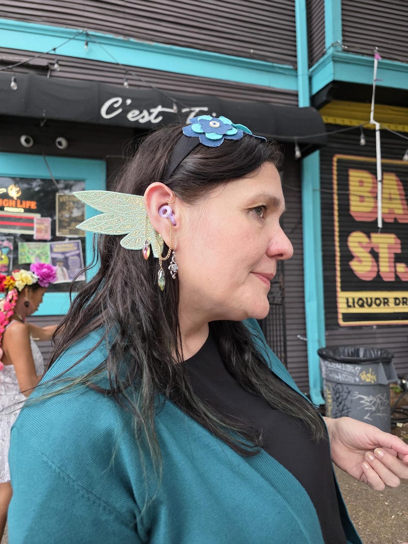 May include: A black headband featuring a layered blue flower ornament, paired with a light green, wing-shaped ear cuff. The cuff includes a delicate gold-toned chain that connects to a dangling, faceted, teardrop-shaped crystal charm.