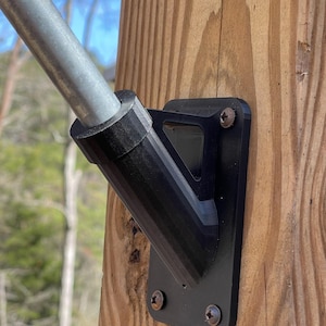 May include: Black metal flagpole bracket mounted on a wooden post. The bracket has a triangular shape and is secured with screws.