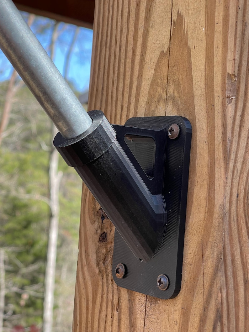 May include: Black metal flagpole bracket mounted on a wooden post. The bracket has a triangular shape and is secured with screws.