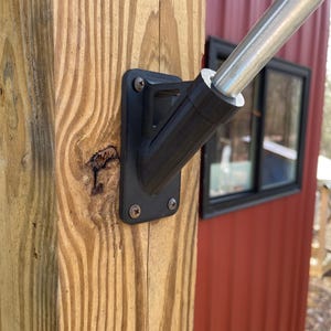 May include: A black metal flagpole bracket mounted to a wooden post. The bracket is attached to a silver flagpole.