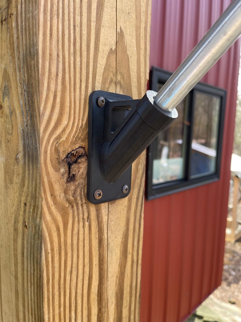 May include: A black metal flagpole bracket mounted to a wooden post. The bracket is attached to a silver flagpole.