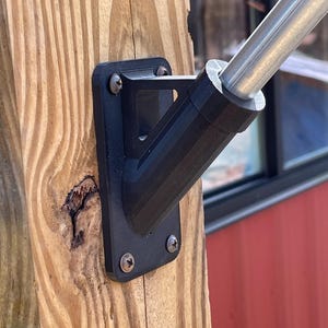 May include: A black plastic flagpole mount with four screws attached to a wooden post. The mount is designed to hold a flagpole at a 90-degree angle.