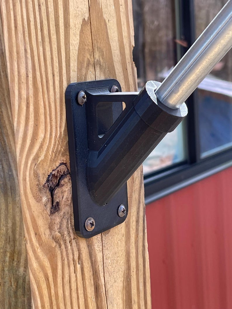 May include: A black plastic flagpole mount with four screws attached to a wooden post. The mount is designed to hold a flagpole at a 90-degree angle.