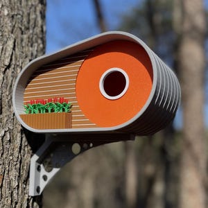 May include: A modern birdhouse with a gray and orange exterior. The birdhouse has a small, round entrance hole and a brown wooden panel with a row of red flowers.