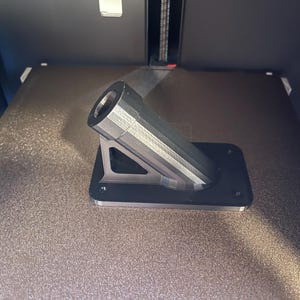 May include: A 3D printed black plastic mount with a cylindrical pipe extending out at an angle. The mount has a triangular shape and is designed to be attached to a surface.