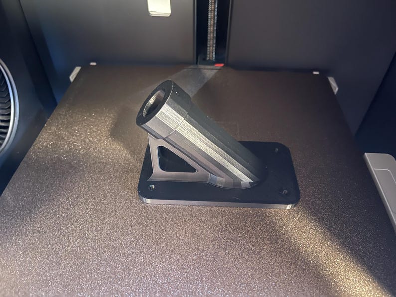 May include: A 3D printed black plastic mount with a cylindrical pipe extending out at an angle. The mount has a triangular shape and is designed to be attached to a surface.