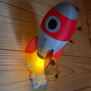 May include: A red, gray, and white rocket-shaped wall light with the name "Piper" printed on the side. The rocket has a glowing yellow light emanating from the base, with white cloud-like details. The light is mounted on a wooden wall.