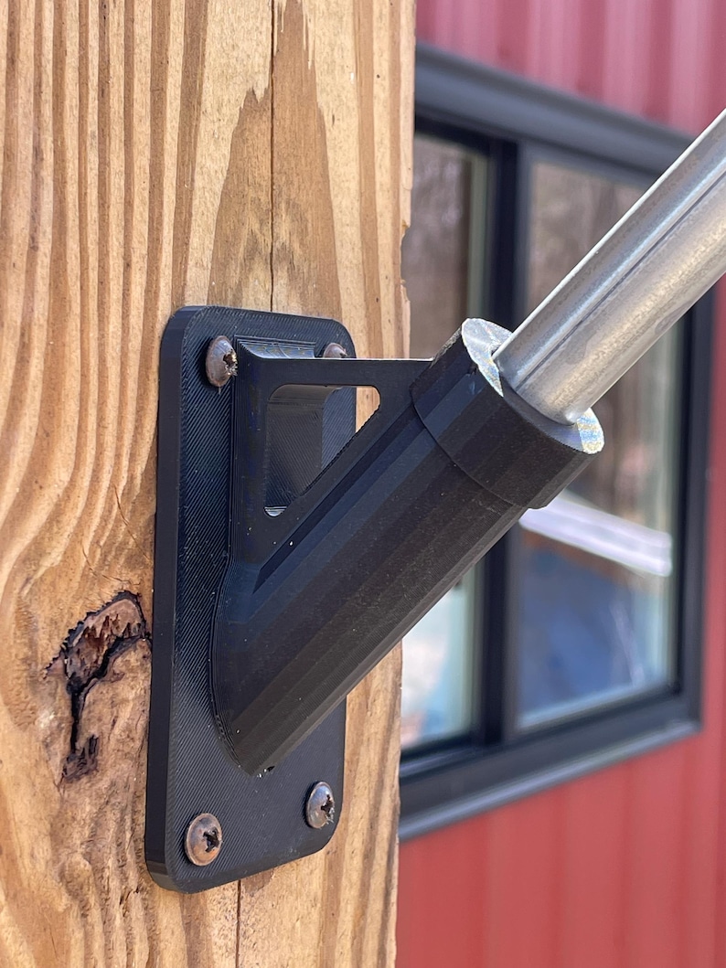 May include: Black plastic flagpole mount with two screws attached to a wooden post. The mount is designed to hold a flagpole at an angle.