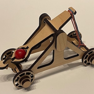 Puede incluir: Un juguete de catapulta de madera con una bola roja en el brazo de lanzamiento. La catapulta tiene una base de madera marrón y detalles negros. Está hecha de madera contrachapada y tiene gomas elásticas para el lanzamiento.