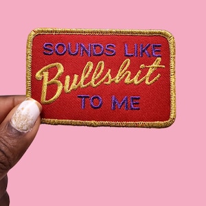 May include: A rectangular red embroidered patch with gold trim and the phrase "SOUNDS LIKE Bullshit TO ME" in gold and purple thread. The patch is held against a pink background.