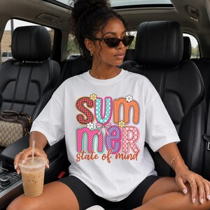 Pode incluir: Uma t-shirt branca de manga curta com o texto colorido "SUMMER" em letras grandes e estampadas, decorado com pequenas flores e um laço, seguido pelo texto laranja em cursivo "state of mind" impresso na parte da frente.