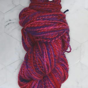 May include: A skein of thick, multi-colored yarn in shades of magenta, red, and purple, twisted into a long oval shape. Two plastic googly eyes are attached to the top section of the yarn, creating a face-like appearance on the fiber bundle.