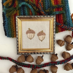 May include: A framed embroidery featuring two acorns with tan and brown thread. The frame is gold with ornate detailing. Acorns and a colorful knitted scarf surround the frame. A red and green knitted garland is at the bottom.