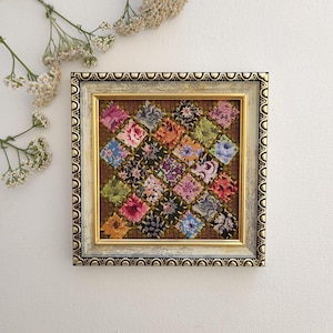 Könnte beinhalten: Quadratisch gerahmtes Kunstwerk mit Patchwork-Design. Das Kunstwerk zeigt ein Gitter aus bunten Stoffquadraten mit floralen Mustern. Der Rahmen hat eine gold-cremefarbene Oberfläche und verleiht dem Stück eine Vintage-Ästhetik.