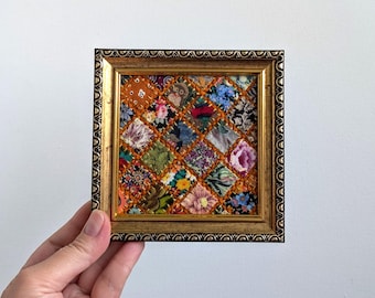 Può includere: Un'opera d'arte da parete quadrata con un patchwork di vari quadrati di tessuto floreale e fantasia disposti a rombi, tenuti insieme da cuciture arancioni, il tutto racchiuso in una cornice dorata decorativa con bordo testurizzato.