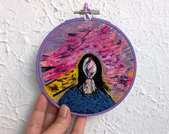 Può includere: Opera d'arte ricamata in un cerchio viola. Il disegno presenta una figura con capelli neri e un top blu, con un volto oscurato da pennellate colorate. Lo sfondo ha sfumature rosa e gialle.