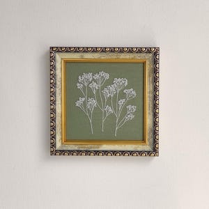 May include: Square framed artwork with white floral embroidery on a sage green background. The frame has a gold inner border and a decorative outer border with black and gold accents. The artwork is displayed against a white wall.