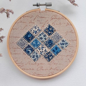 Peut inclure: Un motif de courtepointe patchwork bleu et blanc est cousu sur un fond de tissu beige dans un cercle en bois de broderie. Le tissu a un motif d'écriture manuscrite vintage.