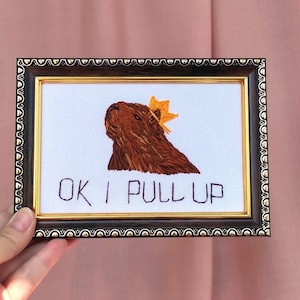May include: Embroidered artwork in a decorative frame. The image features a brown capybara wearing a yellow crown, with the text "OK I PULL UP" below. The frame has a black and gold ornate design.