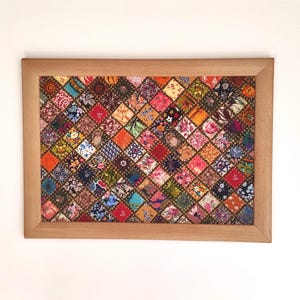 Może przedstawiać: Oprawiony patchwork z jasnobrązową drewnianą ramą. Kołdra składa się z wielu małych, kolorowych kwadratów materiału, każdy z unikalnym kwiatowym lub wzorzystym wzorem. Paleta kolorów obejmuje czerwienie, pomarańcze, błękity i zielenie.
