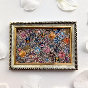 Op de afbeelding: Een ingelijst textielkunstwerk met een patchwork-ontwerp met diverse kleurrijke bloempatronen. De lijst heeft een gouden binnenrand en een verweerde, gebroken witte en gouden buitenrand. Witte bloemblaadjes omringen het kunstwerk.