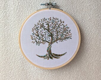 以下が含まれることがあります： 木製の円形刺繍枠に白い布が張られ、手縫いで木の模様が施されています。木はねじれた幹と枝を持ち、緑、茶色、金色の多色の糸で刺繍されており、細部まで描き込まれた樹冠が表現されています。