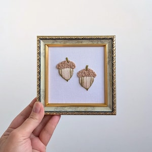 May include: A square decorative frame with a gold and mottled grey finish containing white fabric embroidered with two acorns. Each acorn features a textured, light brown cap and a cream-coloured base with vertical stitching and dark green stems.