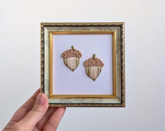 Può includere: Una cornice decorativa quadrata con finitura dorata e grigia maculata contenente tessuto bianco ricamato con due ghiande. Ogni ghianda presenta un cappuccio testurizzato marrone chiaro e una base color crema con cuciture verticali e steli verde scuro.
