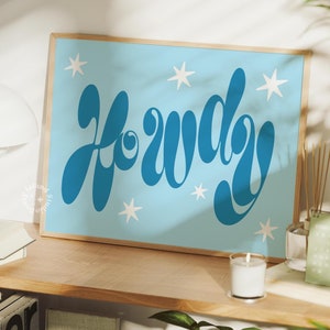 May include: A framed print with a light blue background and white stars. The print features the word "Howdy" in a blue, stylised font.